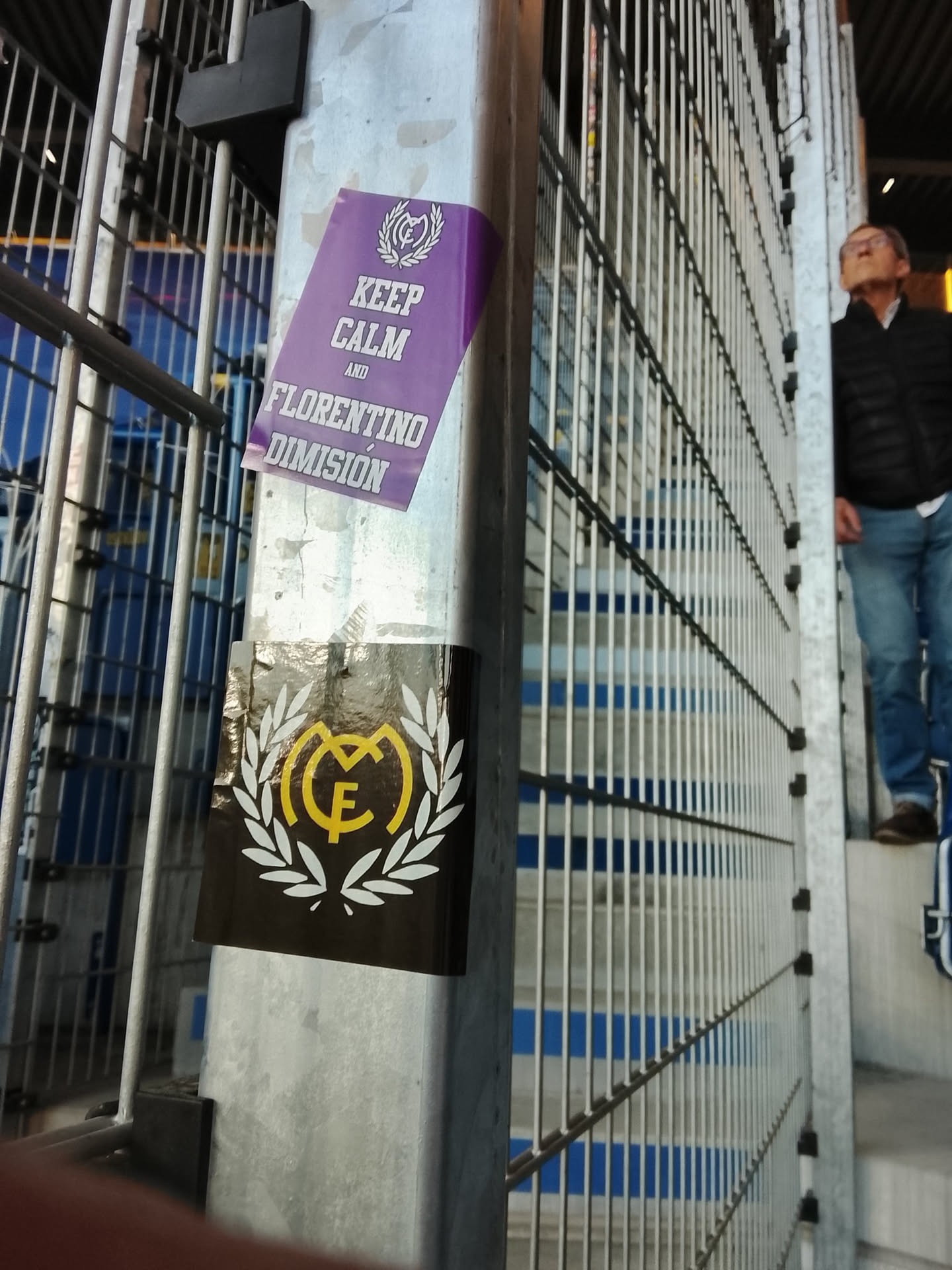 Two rectangular stickers are affixed to a grey metal pole within what appears to be a stadium or sports venue. The upper sticker is predominantly purple with white text and imagery. It features a stylized white crest at the top, resembling a football club's emblem with a crown, intertwined letters (likely 'M', 'C', 'F' for Madrid Club de Fútbol), and a surrounding wreath. Below the crest, the text 'KEEP CALM AND FLORENTINO DIMISIÓN' is printed in a bold, sans-serif, uppercase white font. This sticker shows slight peeling at its top right corner. Directly beneath it, a second, smaller rectangular sticker with a black background is visible. This sticker displays a prominent, stylized yellow 'CF' monogram, encircled by a white laurel wreath.