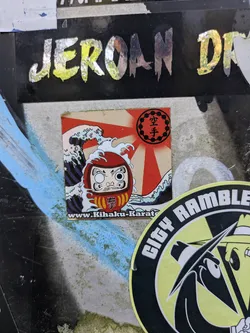The central sticker depicts a red Daruma doll with one eye filled in, set against a dynamic background of stylized blue and white ocean waves and a prominent red rising sun (Hinomaru). Above the Daruma, within the sun's rays, the Japanese characters for 'Karate' (空手) are displayed inside a black circular border. On the Daruma's body, another Japanese character, '拳' (ken, meaning fist), is visible. Along the bottom edge of the sticker, a website address 'www.Kihaku-Karate.com' is printed.