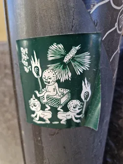 A dark green sticker features an illustration of strange, almost skeletal creatures. Two small figures pull a matchbox with a larger, seated figure atop it. All figures seem to be carrying torches, and a stylized bird is seen near the top of the illustration.  The text on the sticker is difficult to fully make out but appears to be in vertical script.