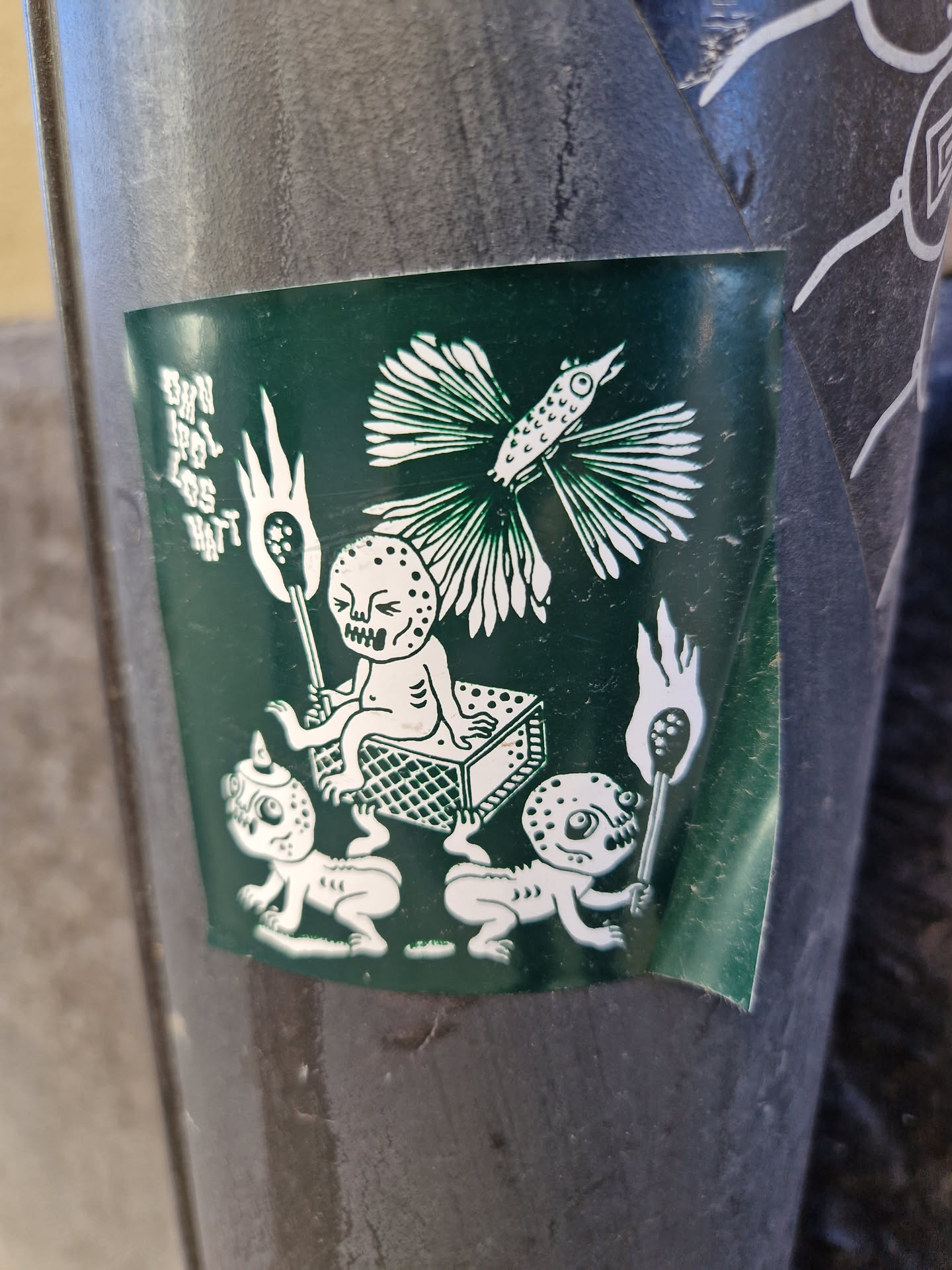 A dark green sticker features an illustration of strange, almost skeletal creatures. Two small figures pull a matchbox with a larger, seated figure atop it. All figures seem to be carrying torches, and a stylized bird is seen near the top of the illustration.  The text on the sticker is difficult to fully make out but appears to be in vertical script.