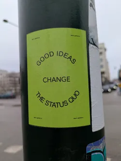 This image features a green rectangular sticker adhered to a pole. The sticker contains the message "GOOD IDEAS CHANGE THE STATUS QUO" in bold, black sans-serif font. The text curves to give it a circular form. There are some inscriptions written in a small font that reads "aon gallery".