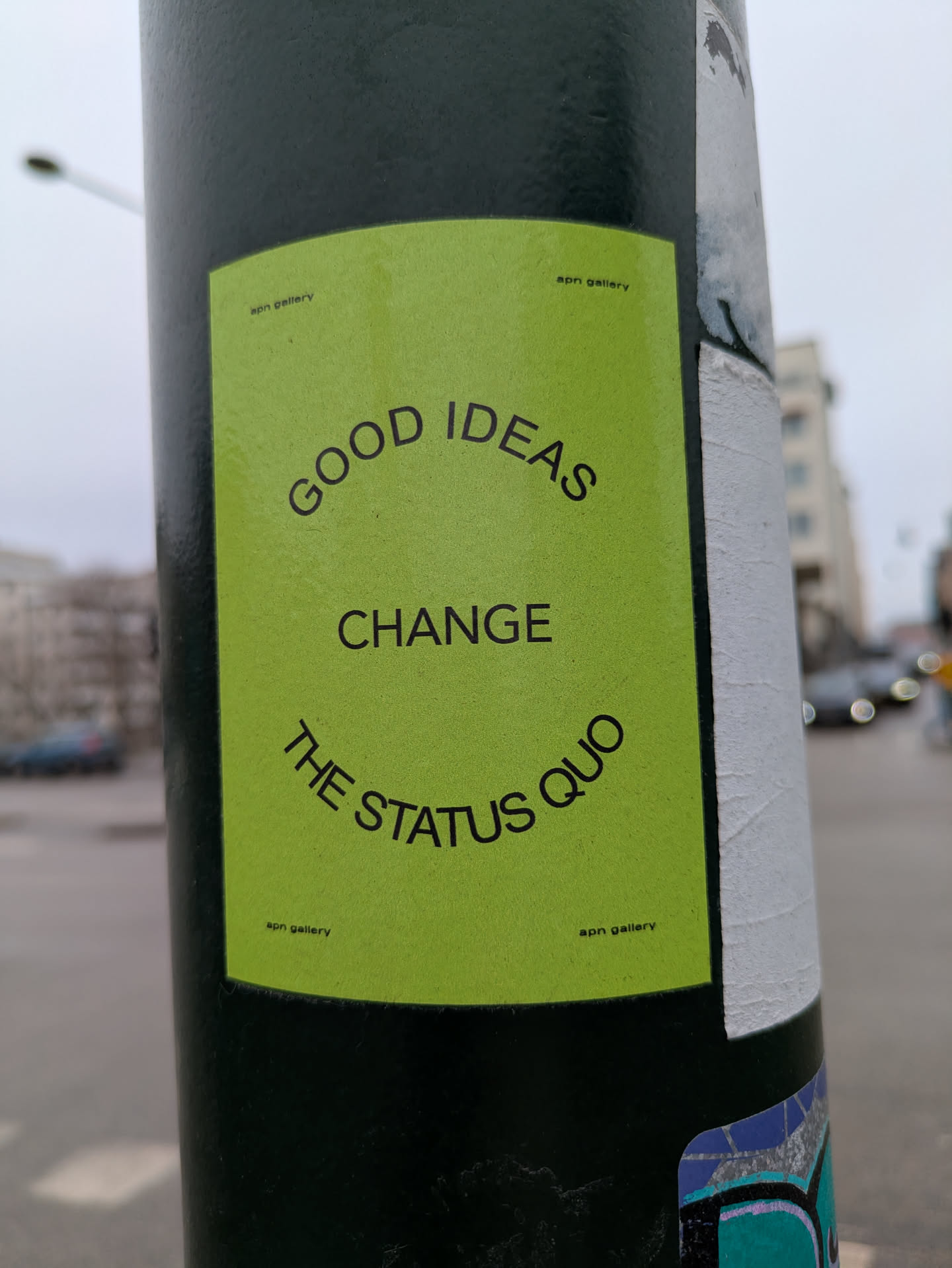 This image features a green rectangular sticker adhered to a pole. The sticker contains the message "GOOD IDEAS CHANGE THE STATUS QUO" in bold, black sans-serif font. The text curves to give it a circular form. There are some inscriptions written in a small font that reads "aon gallery".