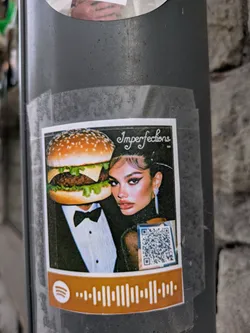 A sticker on a pole features a person wearing a tuxedo with a hamburger head and a woman in a black dress. The sticker has the text 'Imperfections' and includes a Spotify code at the bottom and a QR code next to the woman. The sticker is affixed to the pole with clear tape.
