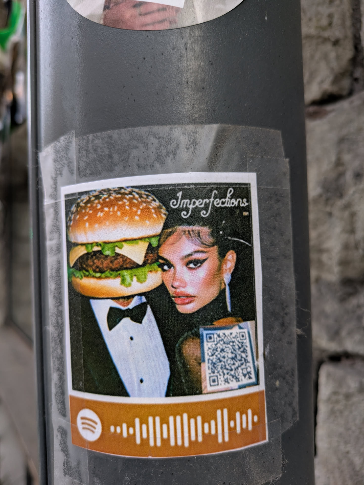 A sticker on a pole features a person wearing a tuxedo with a hamburger head and a woman in a black dress. The sticker has the text 'Imperfections' and includes a Spotify code at the bottom and a QR code next to the woman. The sticker is affixed to the pole with clear tape.