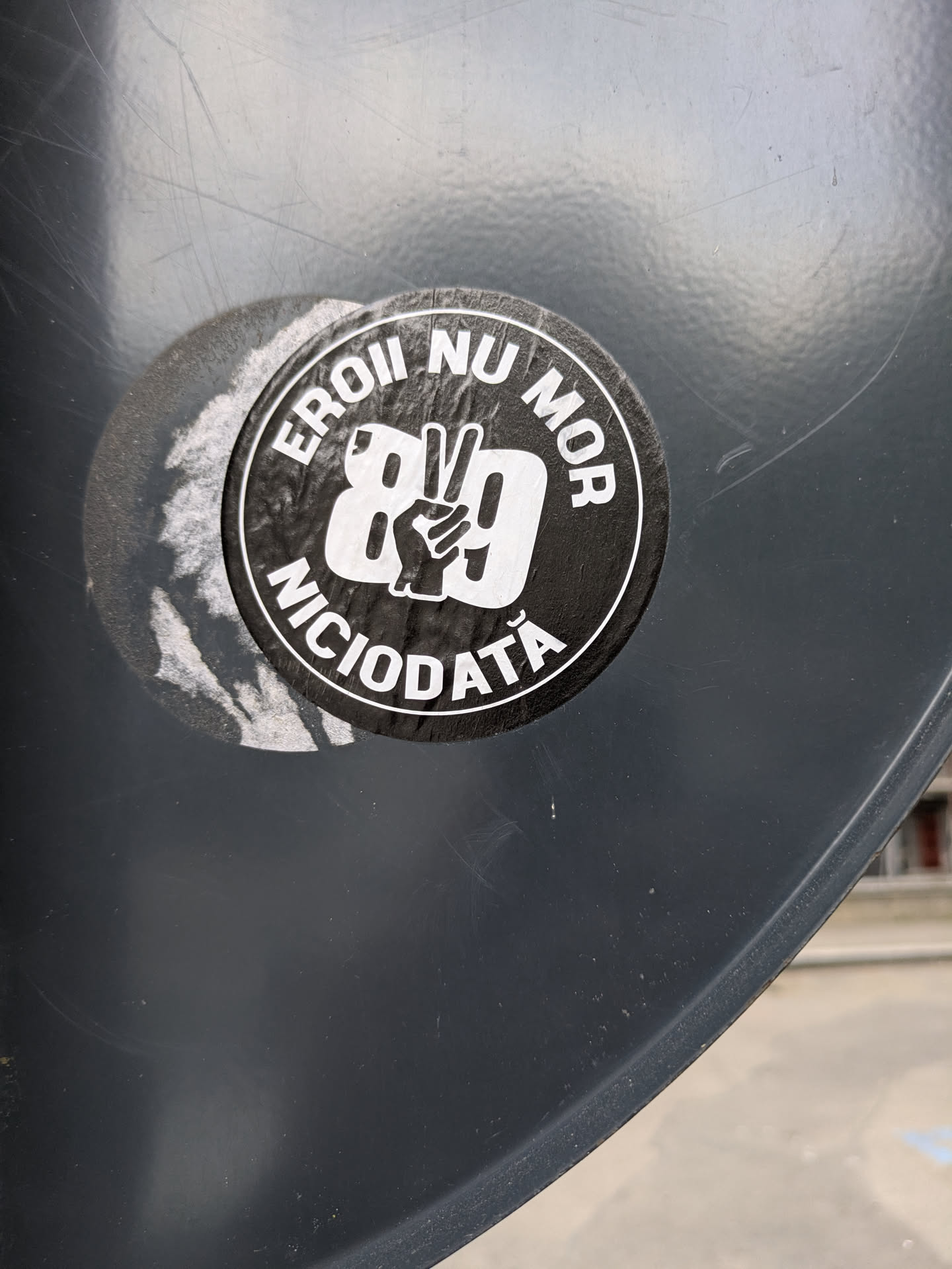 The sticker is on a grey surface. The black sticker has white text around the outer edge and a white peace sign hand gesture over the number 89 in the center. The text reads "EROII NU MOR NICIODATĂ", which translates to "Heroes never die". The sticker appears to be a memorial of the Romanian Revolution of 1989.