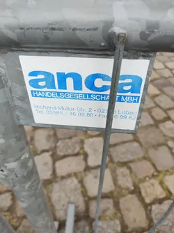 This image shows a sticker on a metal fence. The sticker is white with blue text. The text includes the company name 'anca HANDELSGESELLSCHAFT GMBH', address 'Richard-Müller-Str. 2 02783 Lobau', telephone number '03585/46 89 80', and fax number '6 89 82'.
