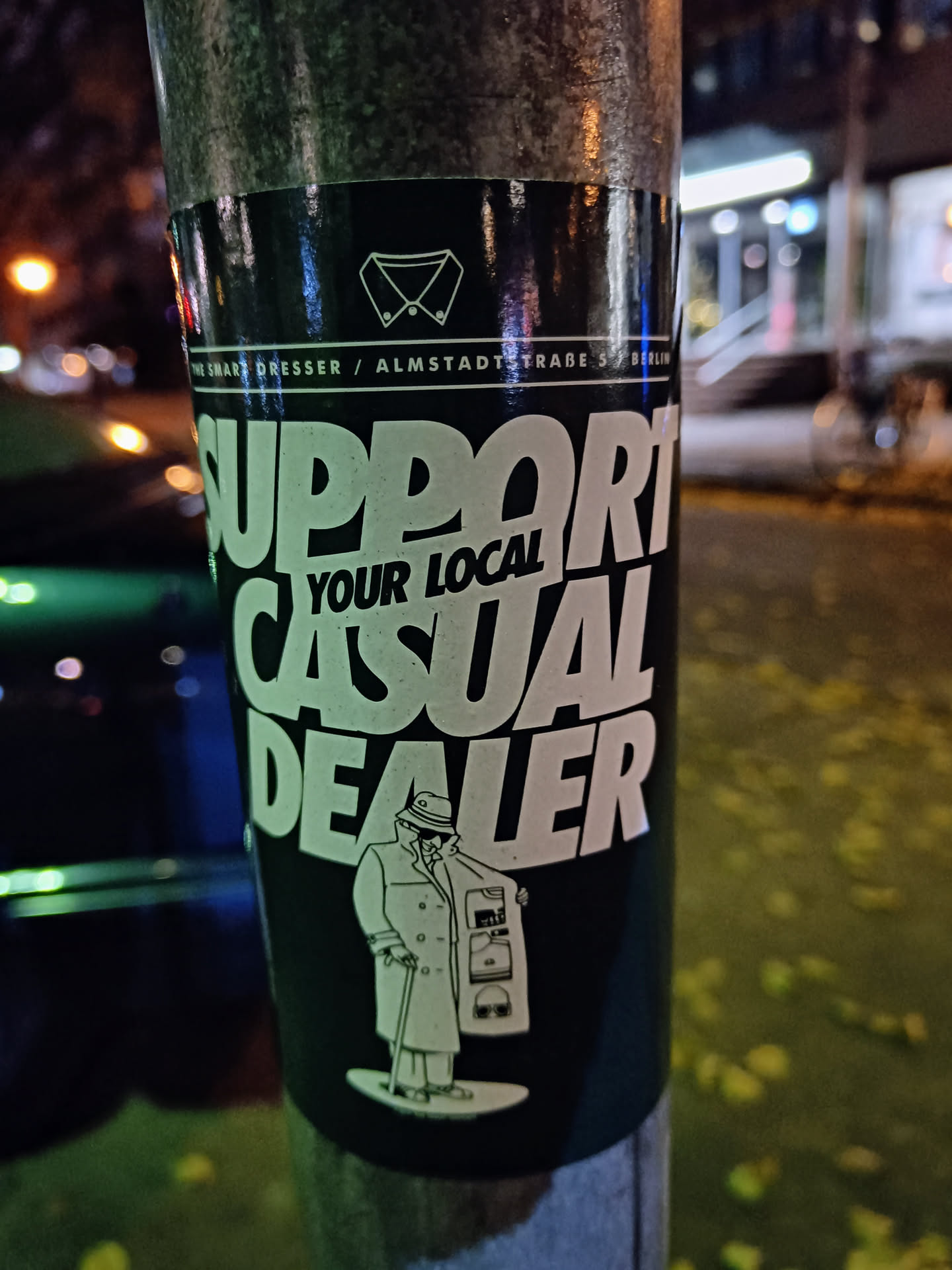 A dark green sticker with white text and an illustration of a man in a long coat holding various items. The text on the sticker says, "Support your local casual dealer." There is also a small logo at the top of the sticker with a shirt collar icon and text which reads "Smart Dresser / Almstadtstraße 57 Berlin".