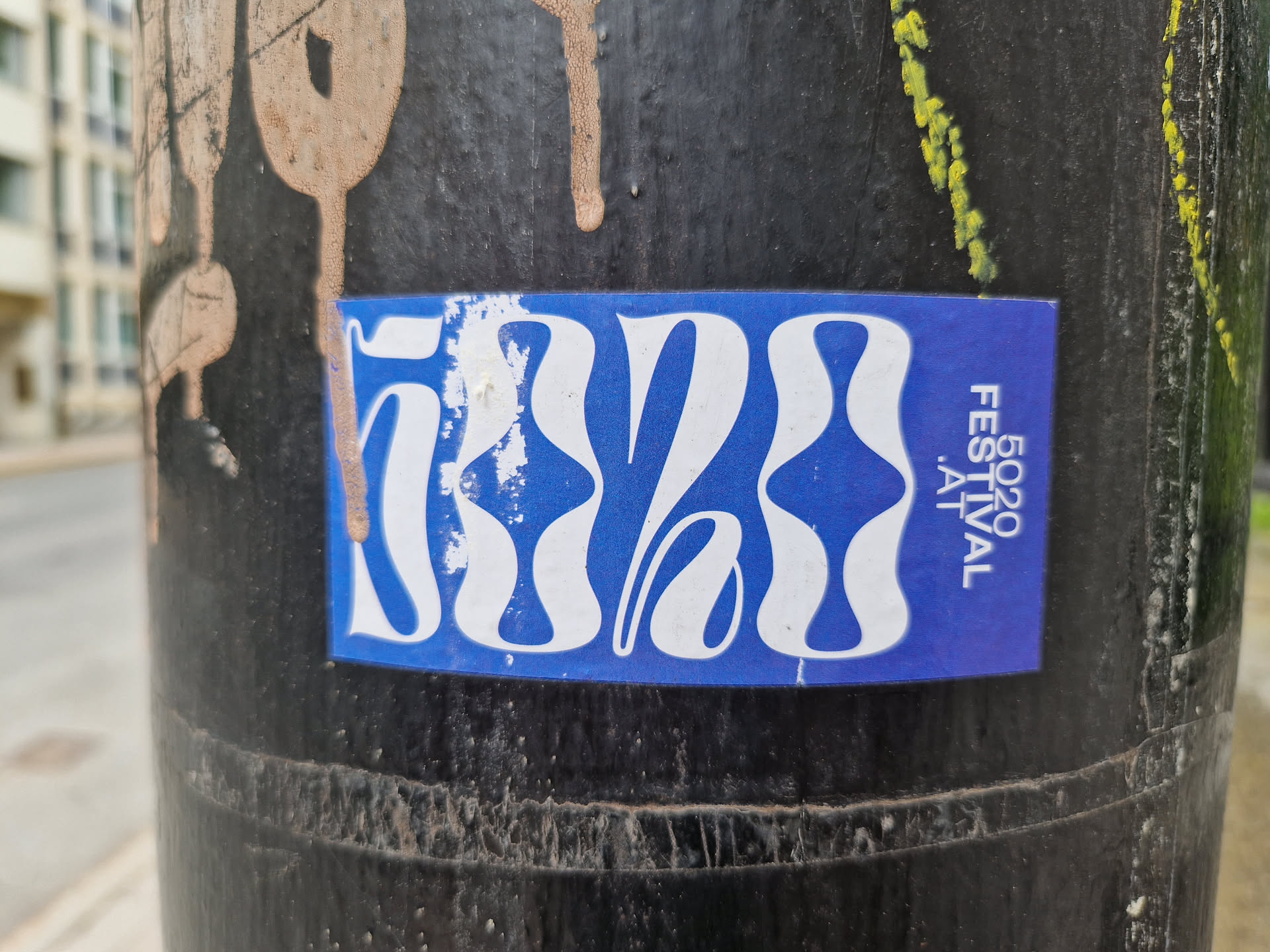 A blue sticker with a white abstract design and the text "5020 FESTIVAL" on it. The sticker is affixed to a dark-colored pole outdoors.