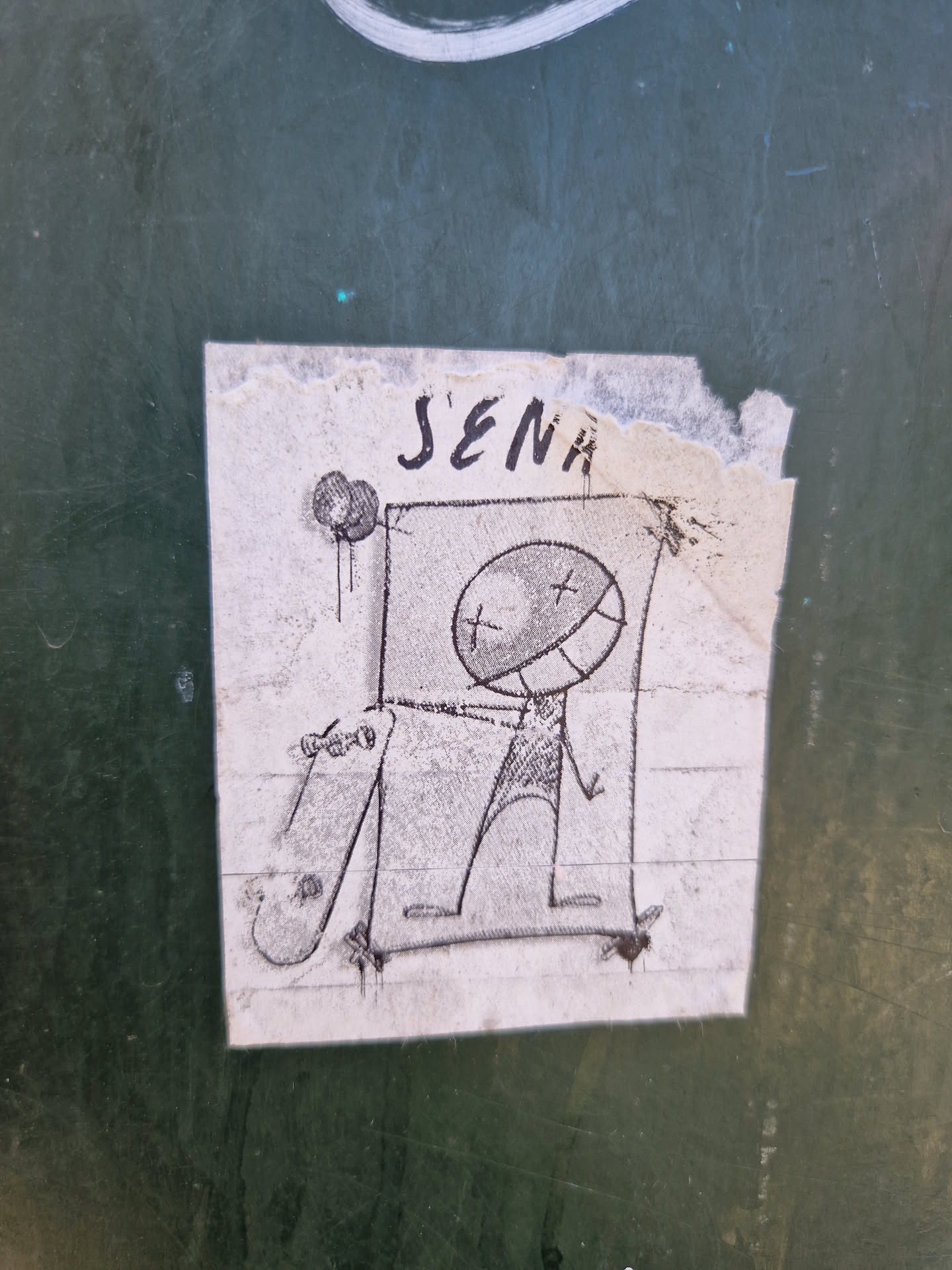 A hand-drawn sticker featuring a simple character design. The character seems to be riding a skateboard. The style is minimalistic, with basic shapes and lines. The sticker has some tears and creases, suggesting it might have been placed outdoors for some time.  The name 'J'ENA' is written at the top.