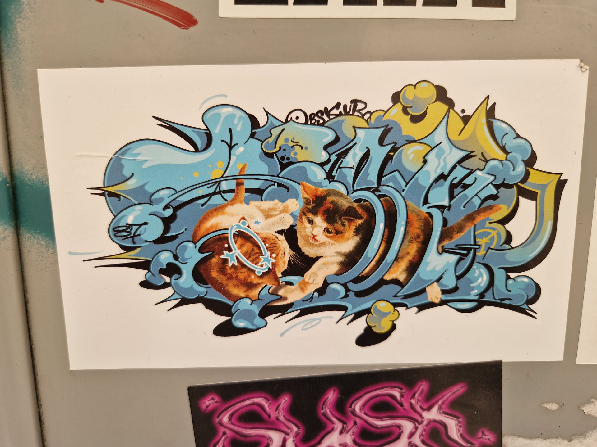 This sticker features a captivating illustration of two kittens interacting within a dynamic graffiti design. The style is bold and playful, with a mix of blues, yellows, and oranges.  The kittens appear to be playfully tumbling within a stylized, abstract form.