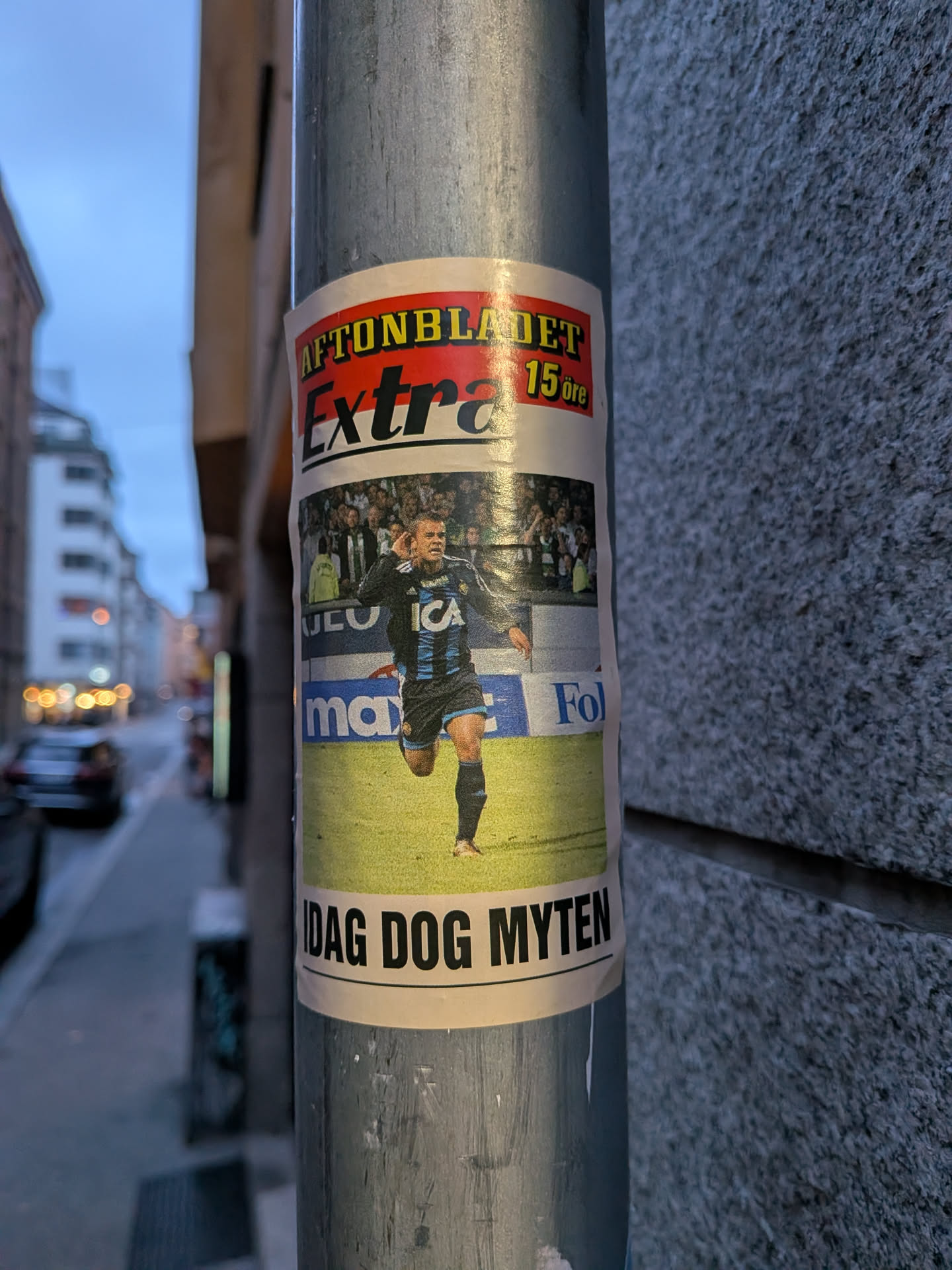 The sticker is styled after a classic Swedish tabloid placard ('löpsedel') from Aftonbladet. At the top, a yellow banner features the text 'AFTONBLADET' in bold yellow and red lettering, accompanied by 'Extra' in large black cursive and '15 öre' in yellow. Below is a photograph of a professional soccer player dressed in Djurgårdens IF's blue-striped kit with the sponsor 'ICA' across the chest, celebrating a goal on the pitch with his hand to his ear. Under the photo, a bold headline in black capital letters proclaims 'IDAG DOG MYTEN'. The sticker is adhered to a cylindrical metal pole outdoors.