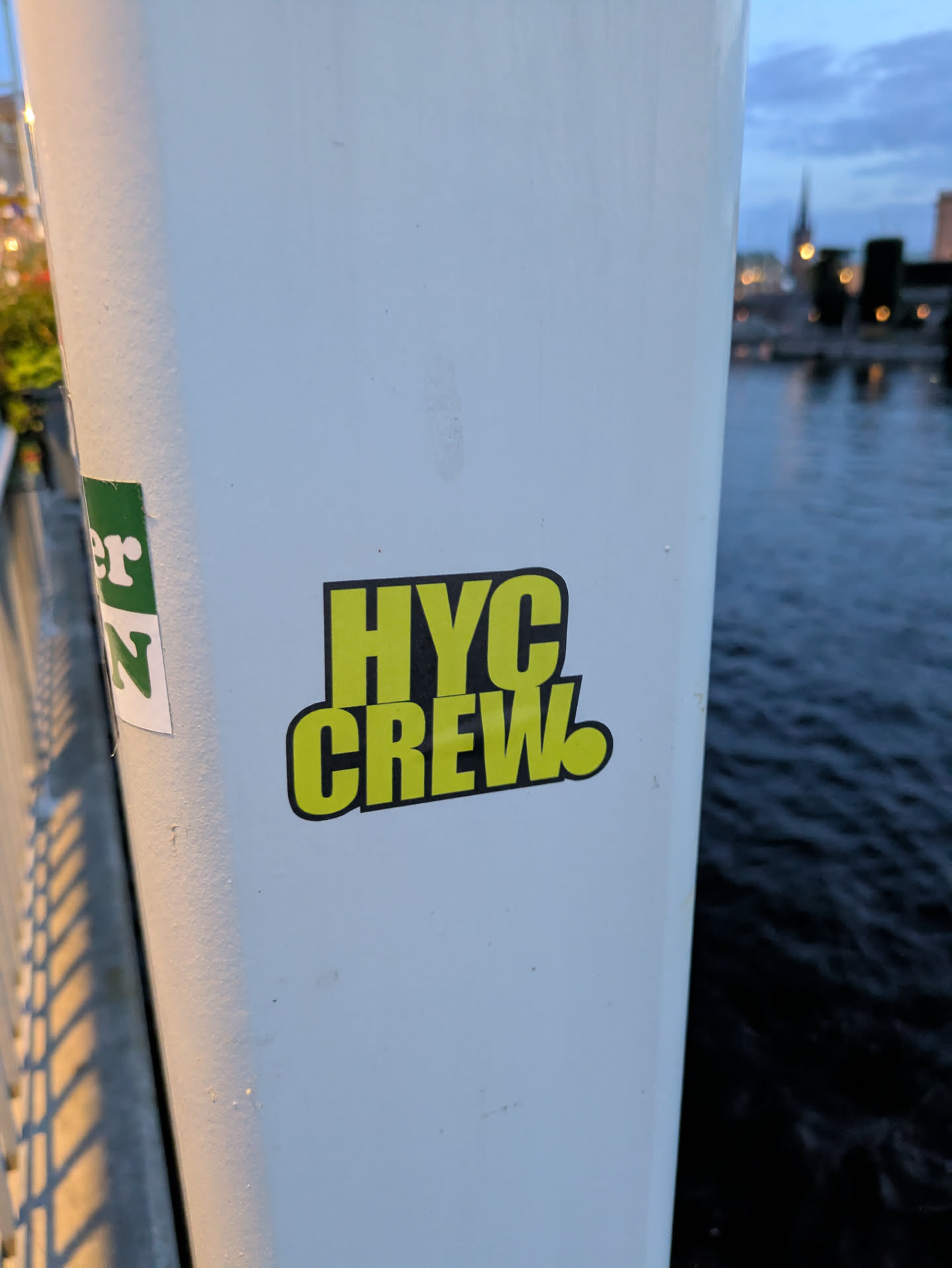 The sticker is centrally located and features the text 'HYC CREW' in large, bold, sans-serif letters. The letters are filled with a bright yellow color and have a thick black outline. The overall shape of the sticker appears to be slightly rounded rectangle. To the left of this sticker, partially visible, is another sticker with green and white elements.