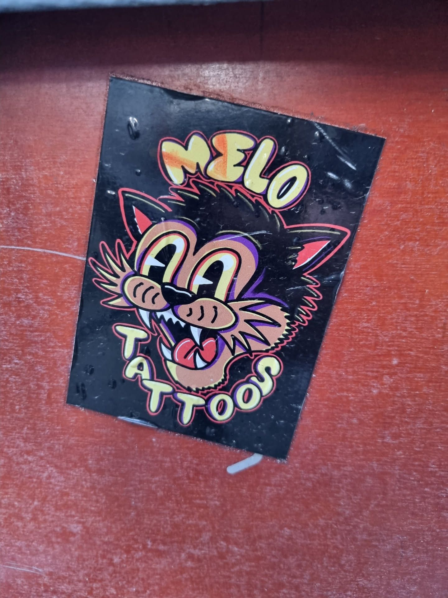 A black sticker with a cartoon cat illustration. The text "MELO" is written above the cat, and "TATTOOS" is written below the cat. The cat's eyes are wide, and it has a large mouth with sharp teeth. The sticker has a slightly worn appearance.