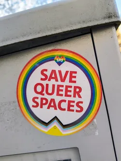A round sticker with a rainbow design and the text "SAVE QUEER SPACES" in red.  A small heart with the letters SPD is in the center of the rainbow. At the bottom is a symbol representing different queer identities.