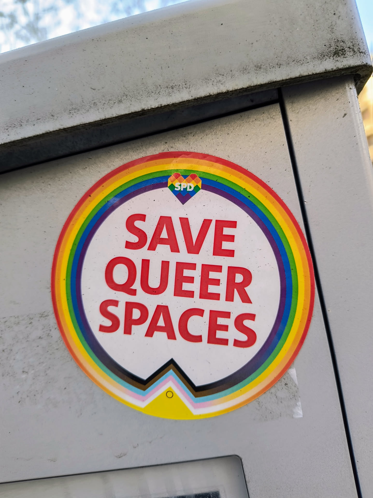 A round sticker with a rainbow design and the text "SAVE QUEER SPACES" in red.  A small heart with the letters SPD is in the center of the rainbow. At the bottom is a symbol representing different queer identities.
