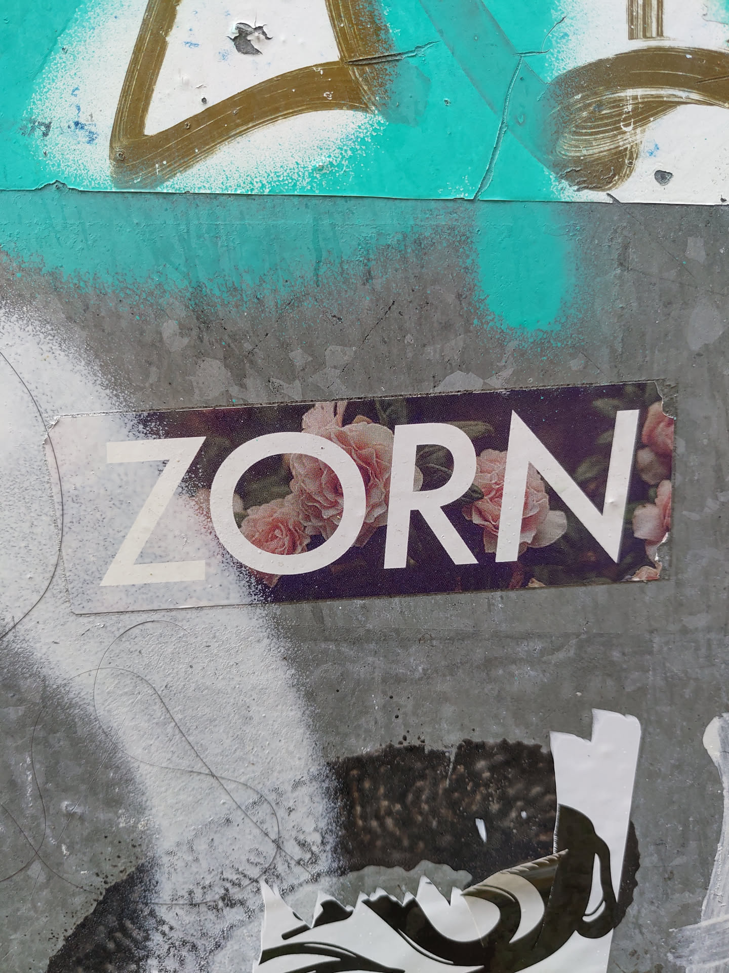 A sticker with the word "ZORN" in white sans-serif font on a background of pink flowers. The sticker is affixed to a grey wall with other graffiti and markings.