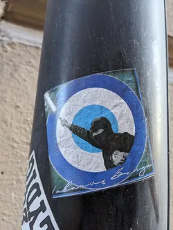 This sticker showcases a unique design of a person in a balaclava throwing something against a blue and white circular background. The style suggests a connection to protest movements or activism. There's a handwritten signature at the bottom, which adds an element of authenticity. 
