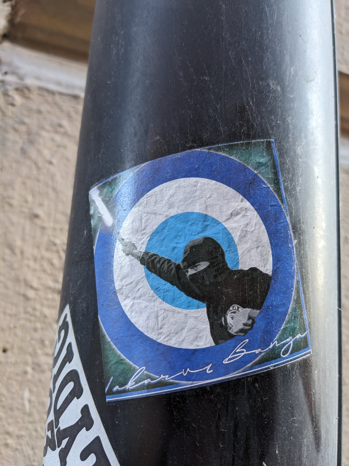 This sticker showcases a unique design of a person in a balaclava throwing something against a blue and white circular background. The style suggests a connection to protest movements or activism. There's a handwritten signature at the bottom, which adds an element of authenticity. 