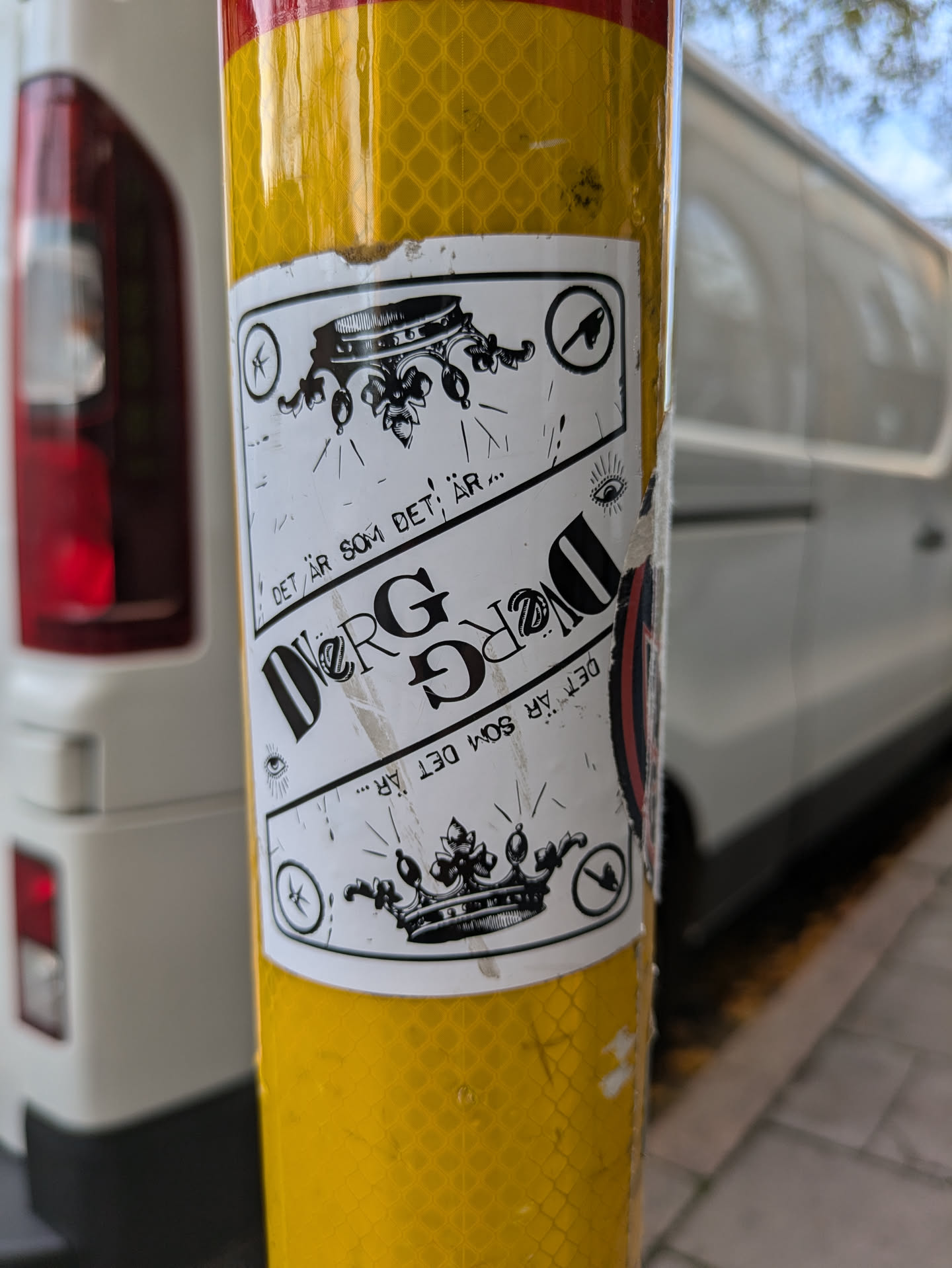 A sticker with a crown and text that says "DET ÄR SOM DET ÄR..." and "...DET ÄR SOM DET ÄR." There is also a symbol that looks like a bird in flight on top of the sticker and a symbol on the bottom that might be a logo. The sticker is attached to a yellow pole.