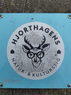 A light blue sign with a white circle in the center. The circle contains a drawing of a deer wearing glasses and the text "Hjorthagens Natur- & Kulturstig".