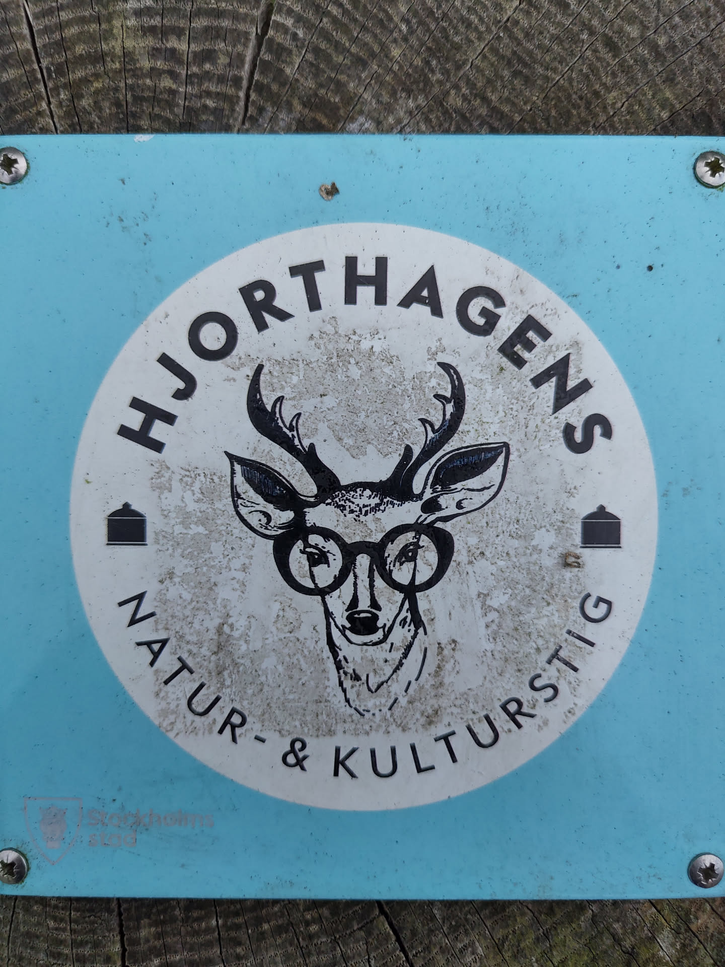 A light blue sign with a white circle in the center. The circle contains a drawing of a deer wearing glasses and the text "Hjorthagens Natur- & Kulturstig".