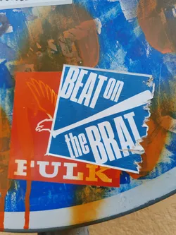 A blue sticker with the text Beat on the Brat in white.  The sticker is slightly torn at the edges and is affixed to a surface with other stickers and markings.