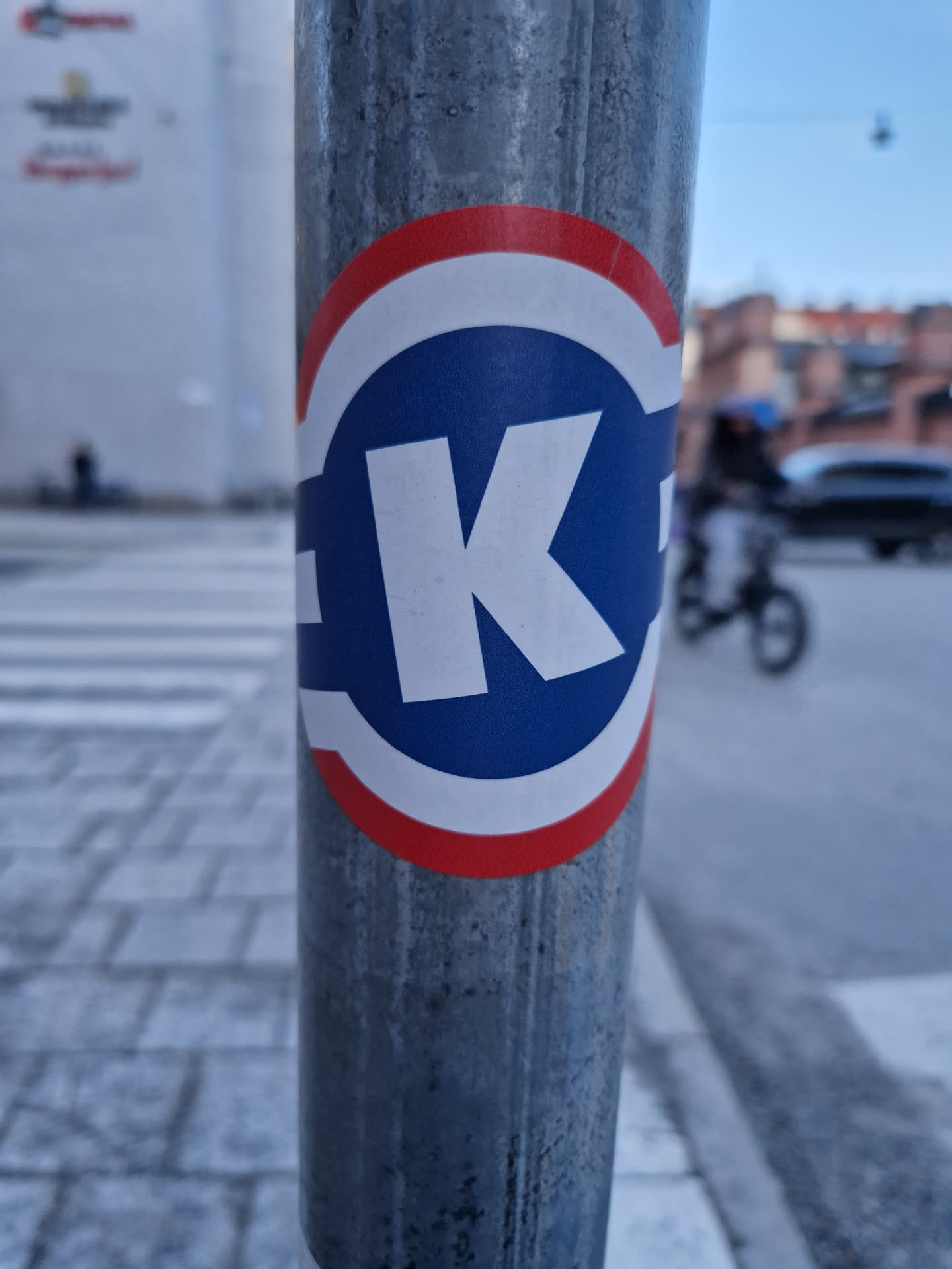 A blue and white circle sticker with a large white "K" in the center. The circle has a red and white border. The sticker is affixed to a grey metal pole, likely outdoors in an urban environment. 