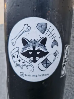 A round sticker featuring a raccoon with hearts for eyes, surrounded by various elements such as bones, a fish skeleton, pizza, and a bottle. The sticker is in black and white and has a vintage tattoo style.  The Instagram handle @trashbaby.tattoos is included at the bottom.