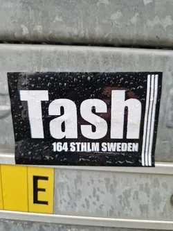 Black and white sticker with the word "Tash" in a large, bold font. Below it, the text "164 STHLM SWEDEN" is printed in a smaller font. The sticker has three vertical lines on the right side and appears to be attached to a metal surface with water droplets visible on its surface.