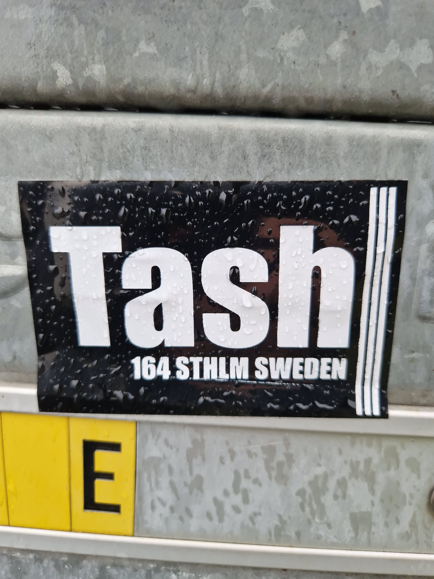 Black and white sticker with the word "Tash" in a large, bold font. Below it, the text "164 STHLM SWEDEN" is printed in a smaller font. The sticker has three vertical lines on the right side and appears to be attached to a metal surface with water droplets visible on its surface.
