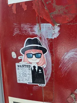 A sticker featuring an anime-style girl in a fedora and sunglasses, holding a "Wanted" poster with a QR code. The style is cute and slightly mischievous. The background is a worn, red surface.