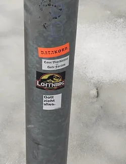 Several stickers are affixed to a gray metal pole.  One sticker says "DATAKORN" in orange text.  Another smaller sticker reads "Love Blackletter - Hate Fascism".  The main sticker features a black and yellow dinosaur with the word "LCHTNBRG" beneath.  A white square sticker below reads "Gott zieht alles."