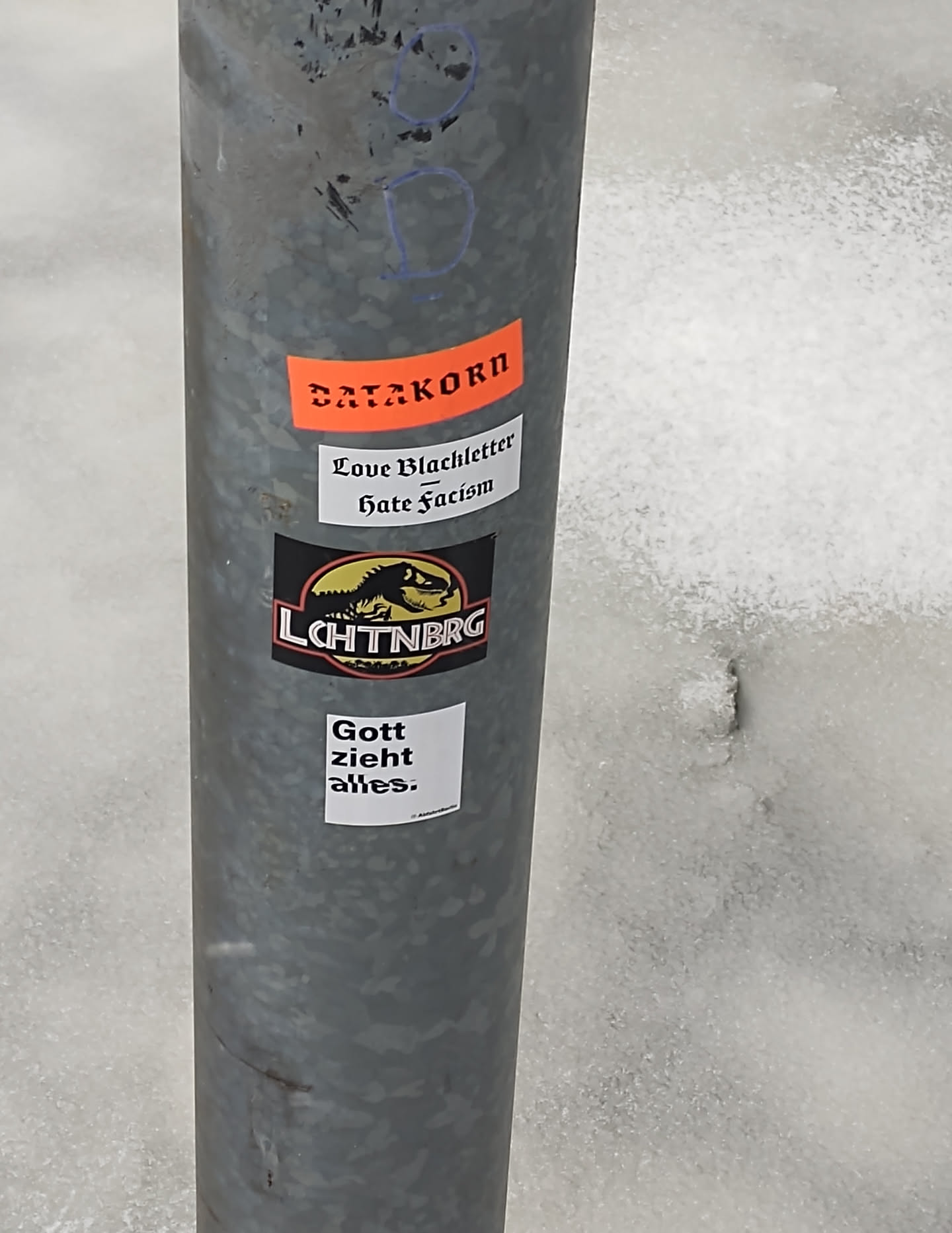 Several stickers are affixed to a gray metal pole.  One sticker says "DATAKORN" in orange text.  Another smaller sticker reads "Love Blackletter - Hate Fascism".  The main sticker features a black and yellow dinosaur with the word "LCHTNBRG" beneath.  A white square sticker below reads "Gott zieht alles."