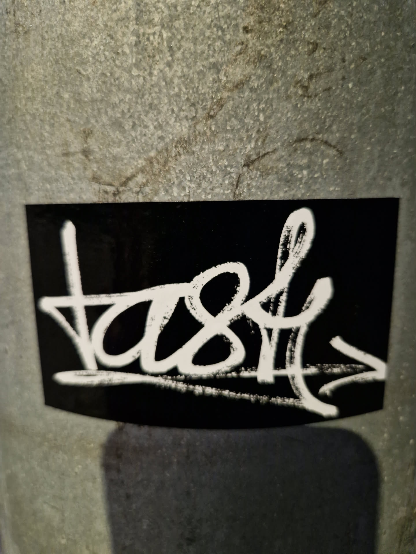 A black sticker with white graffiti-style writing. The word "tast" is written with an arrow pointing to the right.