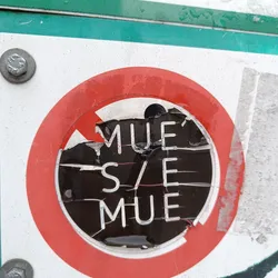 A partially destroyed sticker shows the text 'MUE'S/E MUE' in a stylized font. The sticker is circular with a red border and a diagonal slash across it.  The sticker appears to be affixed to a larger sign or surface, and shows significant wear and tear from its damage.