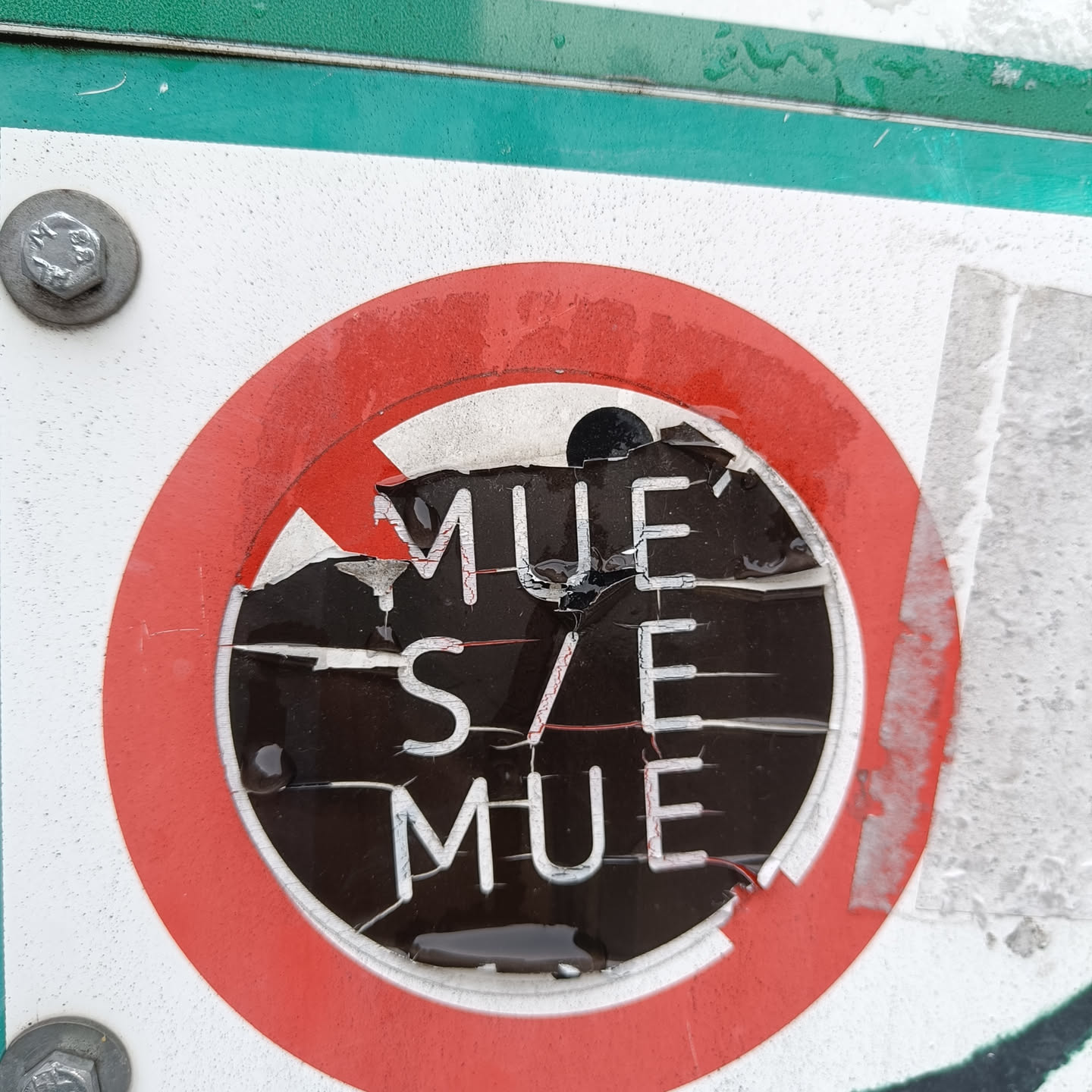A partially destroyed sticker shows the text 'MUE'S/E MUE' in a stylized font. The sticker is circular with a red border and a diagonal slash across it.  The sticker appears to be affixed to a larger sign or surface, and shows significant wear and tear from its damage.