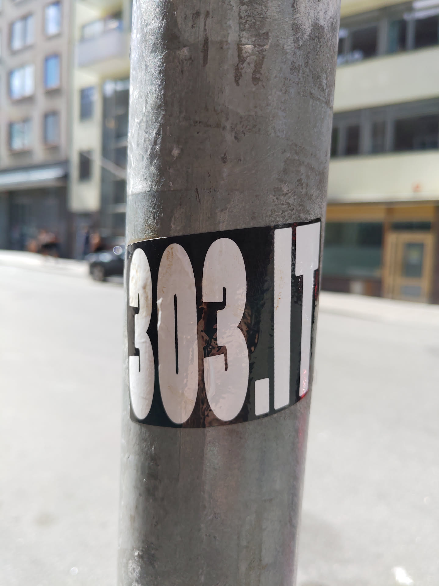A black and white sticker with the text "303.it" in a bold, sans-serif font.  The sticker is affixed to a grey metal pole, likely in an urban setting.  The background is blurry, showing an indistinct city street.