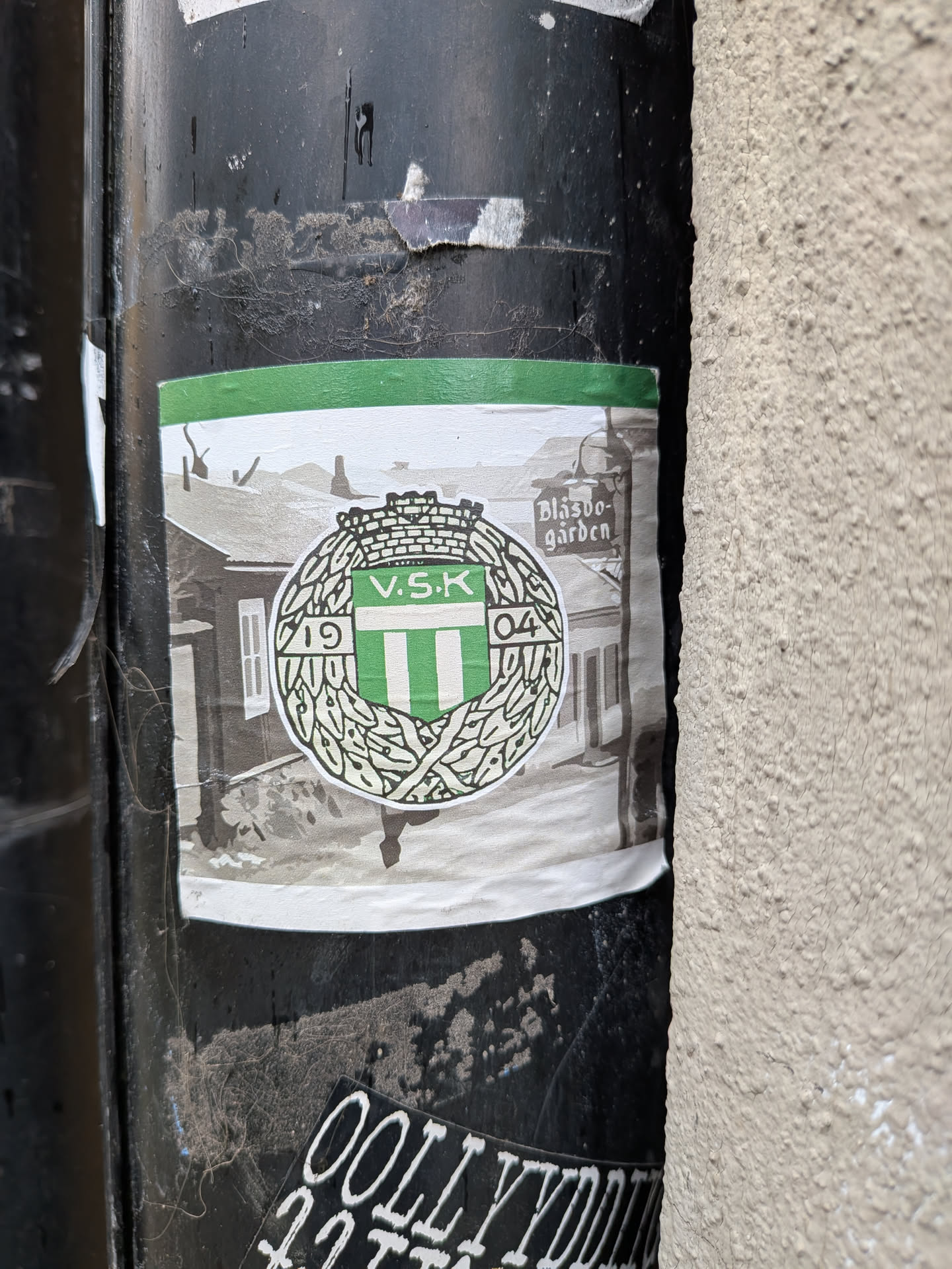 The sticker features a central emblem with a green and white striped shield, topped by a crown, and encircled by a laurel wreath. The letters "V.S.K." are prominently displayed on the shield. To the left of the crest, the number "19" is visible within the wreath, and to the right, "04", likely indicating the year 1904. The crest is set against a grayscale background depicting what appears to be a historical building or street scene, possibly Blåsbo-gården, which is also written on the right side of the background. The top edge of the sticker has a solid green band. The sticker itself is slightly wrinkled and worn, indicating it's been affixed for some time.