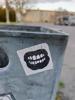 Close-up view of a sticker featuring a detailed drawing of a set of teeth. The sticker has a slightly worn or distressed look, adding to its aesthetic appeal. It is affixed to a grey metallic surface, possibly a bin or container, with some visible wear and tear also visible on the metal.