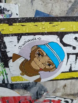 A sticker featuring an illustration of a monkey wearing a blue hat and a gold chain. The style is cartoonish and playful. The sticker appears to be placed outdoors and is wet from rain.