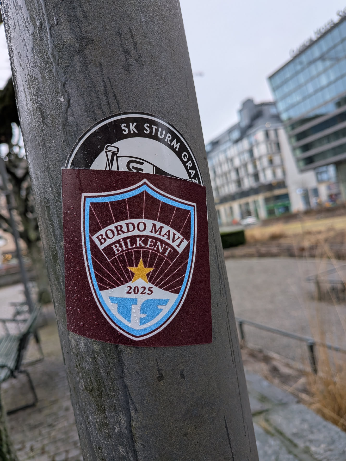 The sticker is a shield-shaped crest, primarily in a deep maroon (bordo) color with light blue and white accents. It features a light blue border around the shield. Inside the shield, the words "BORDO MAVI" are arched at the top, with "BILKENT" directly below them, all in white text. Below the text, a golden-yellow five-pointed star is centered, from which white rays emanate downwards against the maroon background. At the very bottom of the shield, on a light blue band, the year "2025" is written in white, and below that, a stylized 'TS' logo in light blue is set against a white background. The sticker has a slightly glossy finish and shows visible water droplets on its surface, indicating it's exposed to the elements.