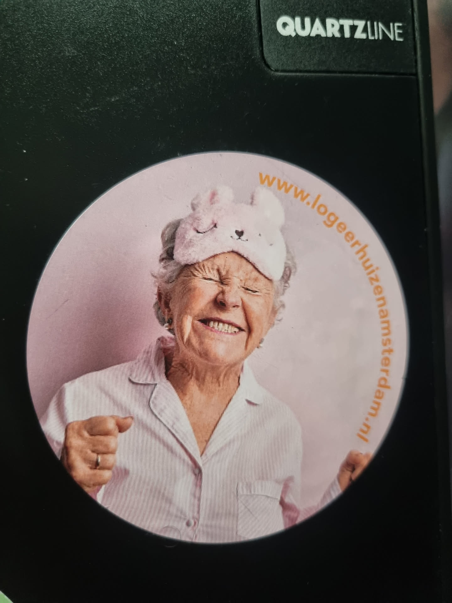 Round sticker featuring a smiling elderly woman wearing a sleep mask.  The background is light pink. Text on the sticker indicates a website address.