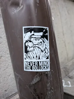 A rectangular, black-and-white sticker affixed to a brown pole. The upper section displays a high-contrast illustration of an aggressive, snarling cartoon character wearing a hat, characterized by a rounded nose, sharp teeth, and a thick, jagged beard. Directly beneath the illustration, bold uppercase text inside a border reads 'NEVER MIND THE BOLLOCKS' arranged in two stacked lines.