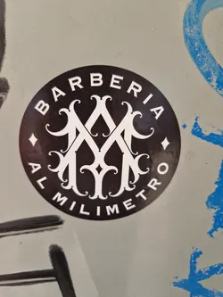 A round black and white sticker with the text BARBERIA AL MILIMETRO and a decorative monogram in the center. The sticker is affixed to a light-grey surface.