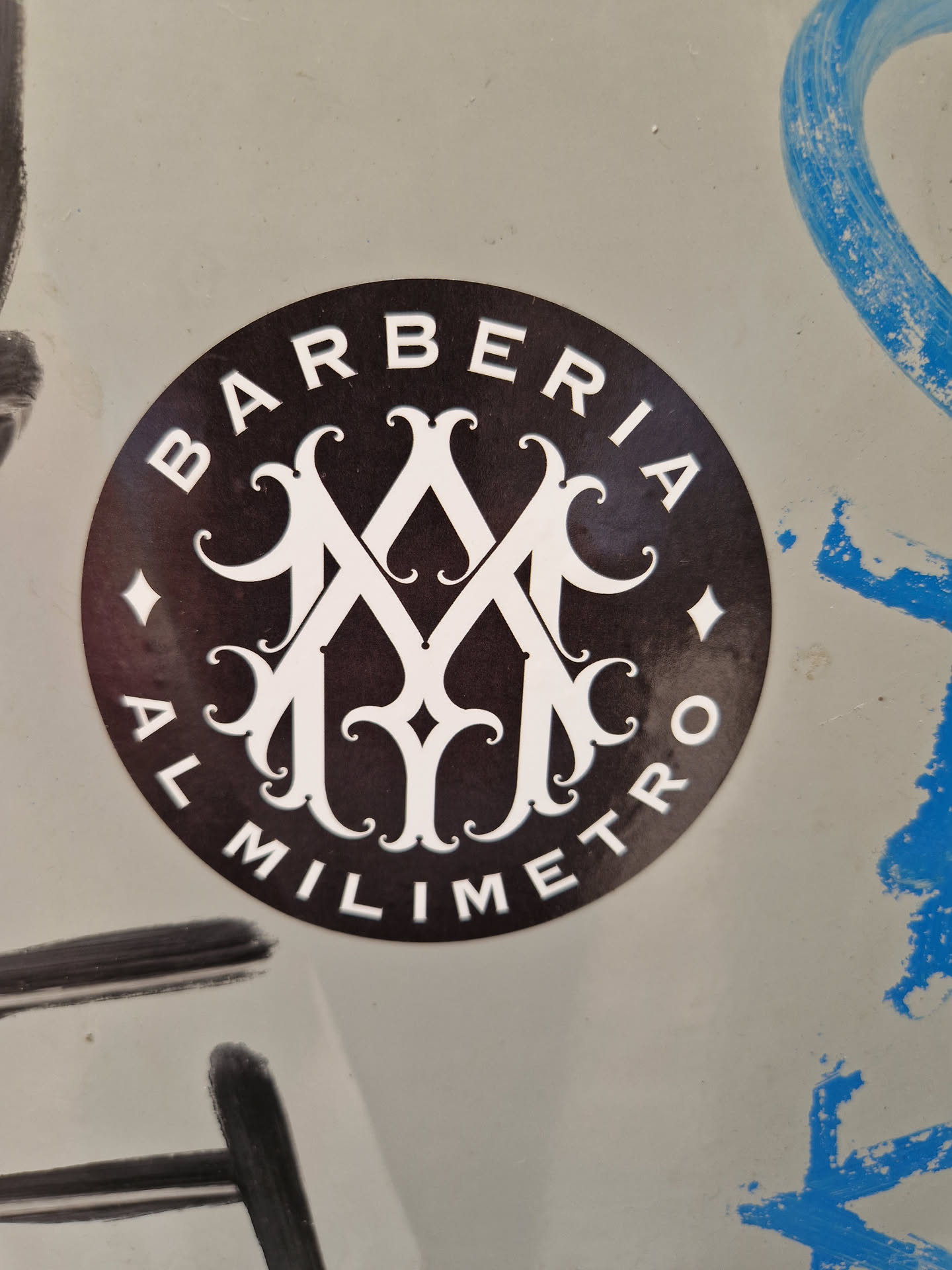 A round black and white sticker with the text BARBERIA AL MILIMETRO and a decorative monogram in the center. The sticker is affixed to a light-grey surface.