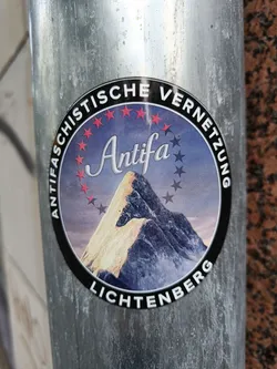 A round sticker with a black border and a picture of a mountain in the center. The text "Antifaschistische Vernetzung Lichtenberg" is written around the sticker. The word "Antifa" is written in a stylized font in the center of the sticker.