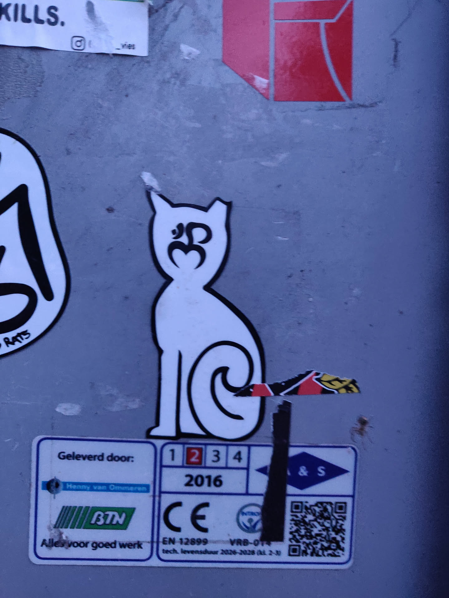 This sticker features a simple line drawing of a cat sitting with its tail curled. There's a symbol resembling a stylized Om symbol on the cat's chest. The sticker is white with black outlines, and it's affixed to a gray surface with other stickers around it. A QR code is visible on a label below the sticker.