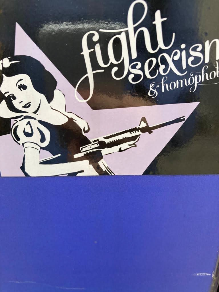 The sticker features a stylized, high-contrast illustration of a character resembling Snow White, rendered in black and white, against a light lilac/pale purple triangular background. The figure is positioned in the lower left, seemingly reclining or leaning, holding an M16-style assault rifle across her body. Her expression is calm and resolute. In the upper right section, on a black background, elegant white script text reads 'fight sexism & homophobia'. The overall aesthetic is graphic and impactful, combining classic pop culture imagery with a strong social message. The sticker itself appears to be affixed to a dark blue surface, which covers the bottom portion of the design.