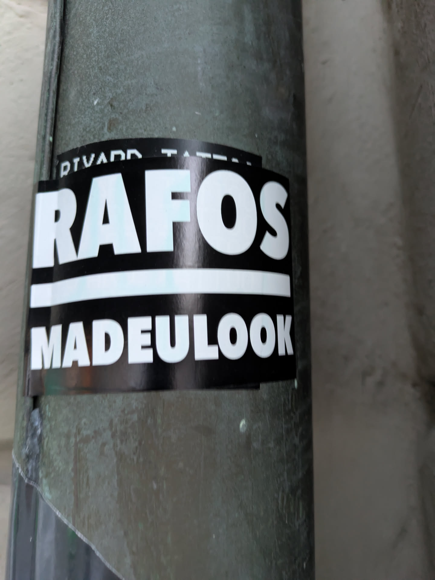 The sticker is predominantly black with white text. The top line of text reads 'RAFOS' in large, bold, sans-serif capital letters with a slight blue or teal outline. Below this is a thin white horizontal line. The bottom line of text reads 'MADEULOOK' in similar bold, white, sans-serif capital letters. The sticker appears to be applied to a cylindrical, greenish-gray surface, possibly a pipe.