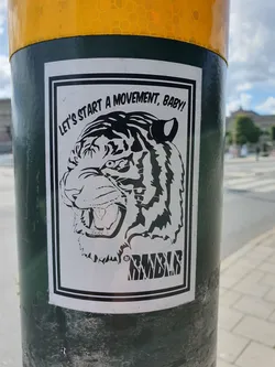 A black and white illustration of a tiger's head with the text "Let's start a movement, baby!" and a logo that says "RMBLR" at the bottom. The sticker is on a dark green pole.